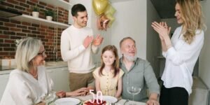 A multi-generational family celebrating a 50th birthday together in a kitchen, featuring an older man, a young girl, and family members clapping around a cake with '50' candles.